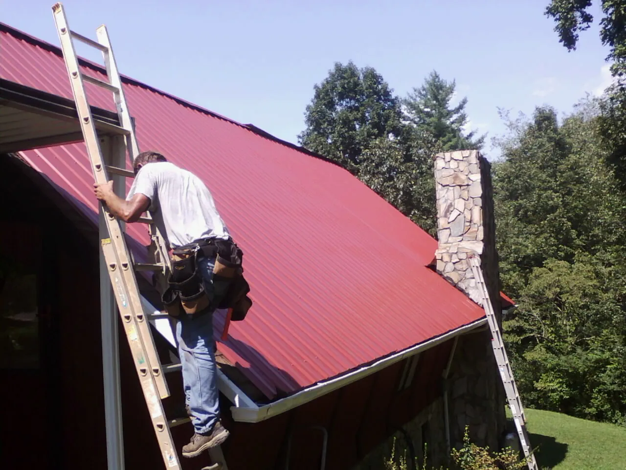 Roofer on a ladder beside a red metal roof and stone chimney — Soffit & Fascia Repair by Roofing Brothers in Keysville